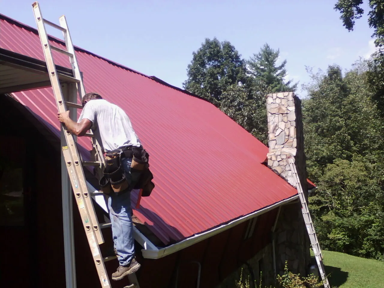 Roofer on a ladder beside a red metal roof and stone chimney — Soffit & Fascia Repair by Roofing Brothers in Randolph